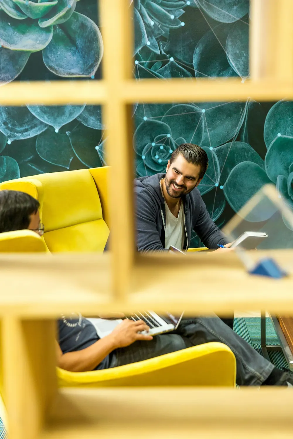 Two people occupy a yellow chair in an office setting, with greenery on the wall behind them.