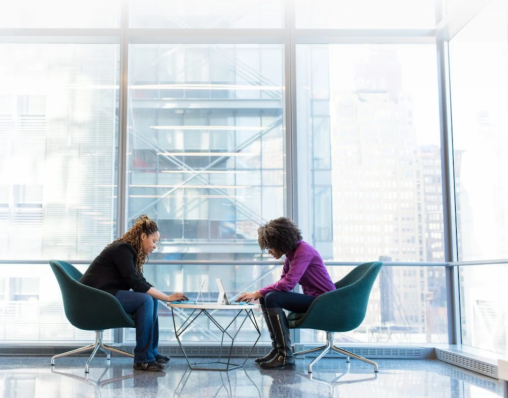 Two women sitting in office chairs, discussing work matters in a professional office environment.