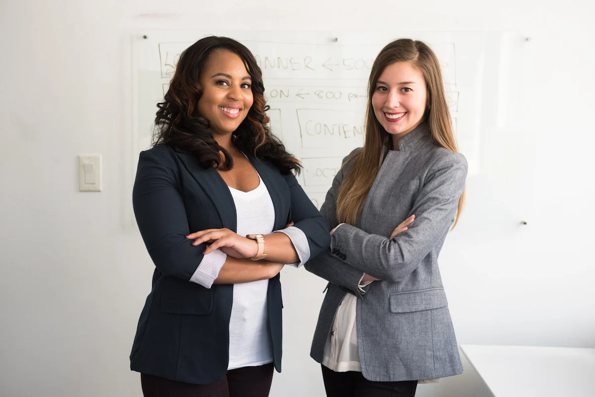 Two women in business attire discussing in front of a whiteboard in a modern office workspace.